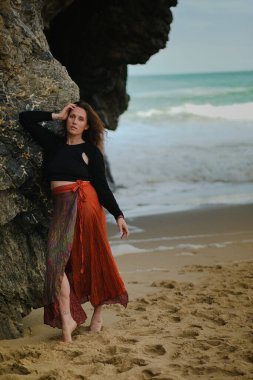Hint tarzı kırmızı etekli güzel bir kadın okyanus kıyısında kayaların yanında poz veriyor. Sahilde pareo giymiş şık bir kız, şık ve şık bir sanat fotoğrafçısı..