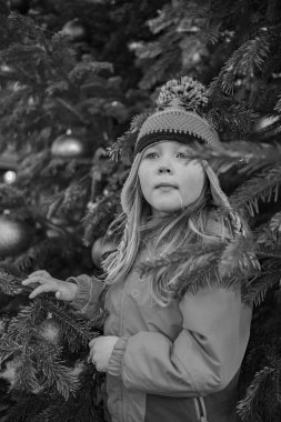 Beş yaşında, kışlık ceketli ve yeni yıl ağacının yanında şapka takan bir kız. Yeni yılda bir kızın portresi.