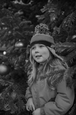 Beş yaşında, kışlık ceketli ve yeni yıl ağacının yanında şapka takan bir kız. Yeni yılda bir kızın portresi.