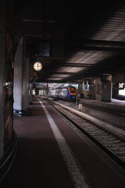 Beton, kasvetli ve karanlık bir tren istasyonu, platformda bir tren duruyor ve duvarda elleri olan bir saat asılı..