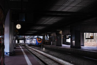 Beton, kasvetli ve karanlık bir tren istasyonu, platformda bir tren duruyor ve duvarda elleri olan bir saat asılı..