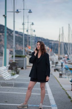 Siyah ceketli ve siyah etekli güzel bir kız bir tatil köyünün arka planına karşı güzel bir sette yürüyor ve konuşuyor..