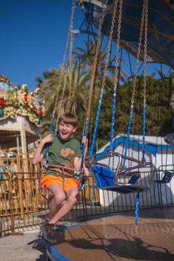Güneşli bir yaz gününde bir çocuk atlıkarıncaya biniyor, gülüyor, el sallıyor ve eğleniyor. Arka planda palmiye ağaçları yetişir ve diğer sürüşler görülebilir..