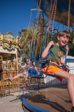 Güneşli bir yaz gününde bir çocuk atlıkarıncaya biniyor, gülüyor, el sallıyor ve eğleniyor. Arka planda palmiye ağaçları yetişir ve diğer sürüşler görülebilir..