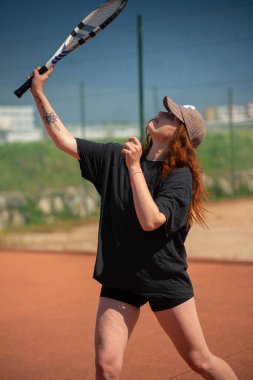 Siyah tişörtlü ve beysbol şapkalı kız güneşli yaz gününde tenis kortunda tenis oynuyor..