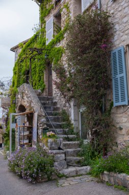 Eski bir kasaba, sarmaşık ve üzümlerle kaplanmış taş bir bina, dar antik bir merdiven girişe açılıyor, mavi panjurlar açık..
