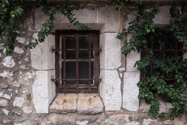 Eski kasaba, taş bina, pencere parmaklıklarla kapatılmış. Paslı dövülmüş parmaklıklar ve duvarlarda büyüyen sarmaşıklar. Eski bir evin penceresi..