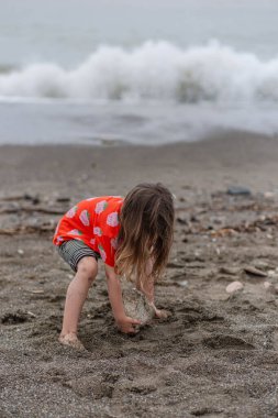 Kırmızı tişörtlü ve çizgili şortlu bir kız kumsalda kum ve taşlarla oynuyor..
