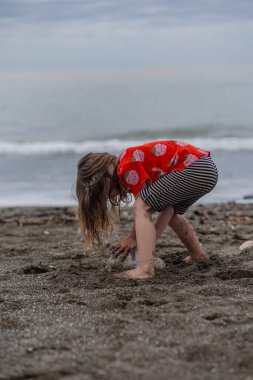 Kırmızı tişörtlü ve çizgili şortlu bir kız kumsalda kum ve taşlarla oynuyor..