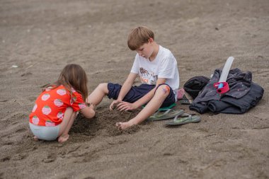 A girl in a red T-shirt and striped shorts plays with her older brother in the sand and stones on the sea beach. Girl and boy on the sea beach on a cloudy day.
