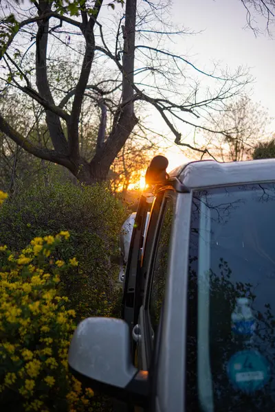 Küçük bir karavan çiçek açan çalıların ve ağaçların arasında duruyor ve batan güneş karavanı aydınlatıyor ve ışınlar doğrudan objektife vuruyor..