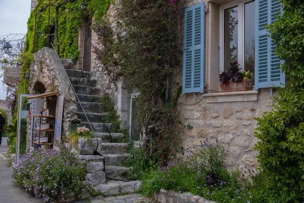 Eski bir kasaba, sarmaşık ve üzümlerle kaplanmış taş bir bina, dar antik bir merdiven girişe açılıyor, mavi panjurlar açık..