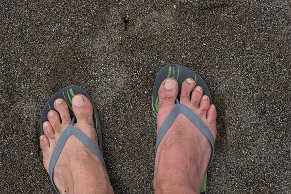 Kumun üzerinde duran lastik parmak arası terlikler içindeki erkek ayakları. Plajda duran lastik parmak arası terlikler, yakın plan fotoğraf..