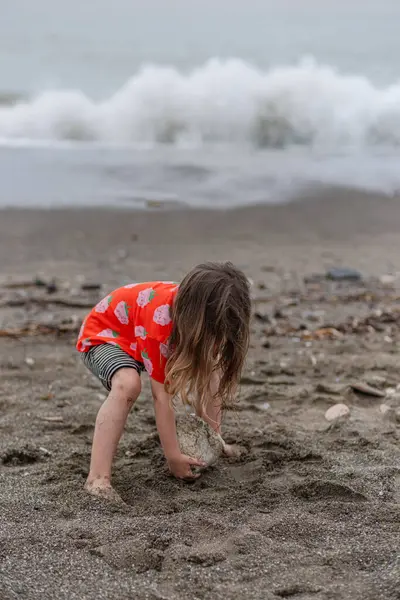 Kırmızı tişörtlü ve çizgili şortlu bir kız kumsalda kum ve taşlarla oynuyor..