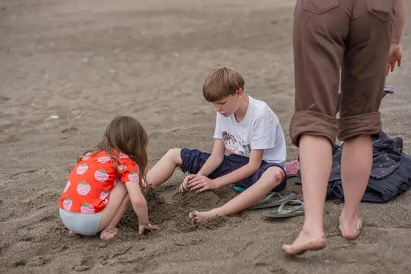 Kırmızı tişörtlü ve çizgili şortlu bir kız, deniz sahilinde kum ve taşlarda ağabeyiyle oynuyor. Kız ve oğlan bulutlu bir günde deniz sahilinde.