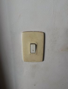 Kabuk soyma duvarında basit ve işlevsel bir tasarımı gösteren bir ışık düğmesi. Düğmenin sarı rengi, aşınma ve yırtık belirtileri gösteren duvarın beyaz zeminiyle çelişiyor..