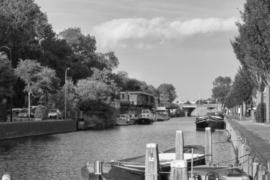 Schwarz-weisse-Aufnahme eines Kanalları, gesaeumt von Baeumen und Gebaeuden. Moderne Wohnungen auf der einen Seite, aeltere Haeuser und Autos auf der anderen. Boote sind entlang der Ufer angedockt.