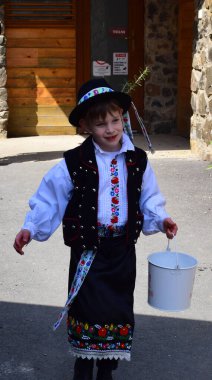Holloko Paskalya Festivali - Holloko, Macaristan - 18 Nisan 2022 - Çocuk Paskalya yağmuru için bir kova su taşıyor. Geleneksel Paskalya halk kostümü giyip bir Paskalya Festivali 'nde kutlama yapmak.. 