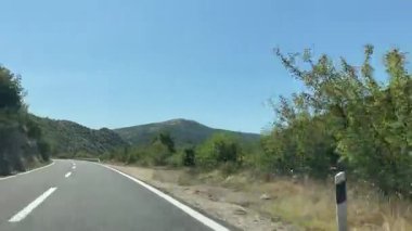 Dashcam POV manzaralı düz, dolambaçlı bir otoyol açık mavi gökyüzünün altındaki yemyeşil tepeleri ve dağları kesiyor. Yol güçlü bir korkulukla kaplı, yavaşça kıvrılıyor ve uzaklarda kayboluyor. Açık yol seyahatleri, araba yolculukları..