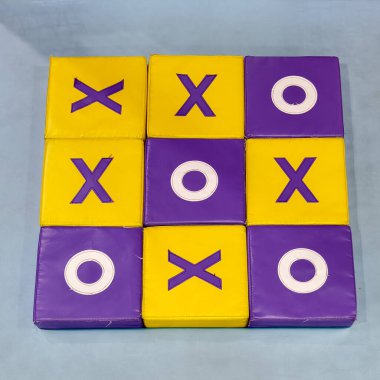 Sarı ve mor renkli yumuşak bloklardan oluşan canlı bir Tic Tac Toe oyun tahtası, kabartmalı X ve O sembolleri oyuncu bir desen içeriyor