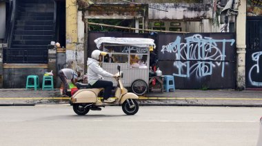 Sukabumi, Endonezya - 3 Mart 2025: Bir adam, kentin bir bölgesinde, duvar yazıları ve yıpranmış bir binanın yer aldığı bir sokak yiyecek arabasının önünden klasik scooter ile geçiyor