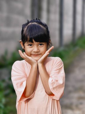 Pembe tişörtlü çekici bir genç kız, ışıl ışıl gülümsüyor, açık havada yemyeşil bir ortam içinde.