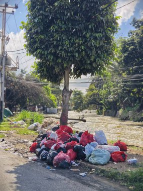 Bogor, Endonezya - 14 Nisan 2025: Nehir ve cadde yakınlarındaki bir ağacın yanına yığılmış çöp torbaları çevresel bir endişe yaratıyor.