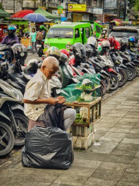 Bogor, Endonezya - 20 Nisan 2025: Yaşlı bir adam kalabalık bir motosiklet parkının yanında elinde tavşan kafesiyle kaldırımda oturuyor