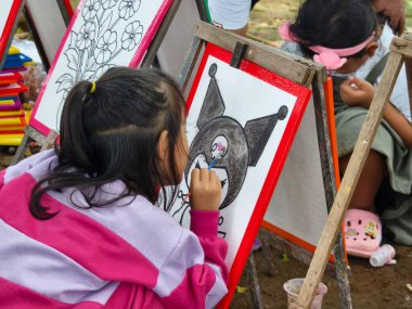 Bogor, Endonezya - 20 Nisan 2025: Genç bir kız tuvale bir karakter çizerken, diğer bir çocuk açık hava halk etkinliğinde çiçek çiziyor. 