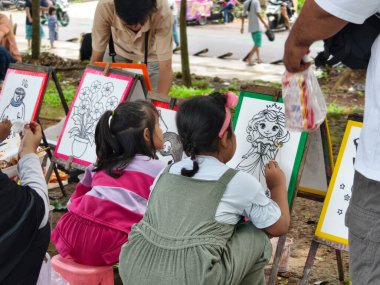 Bogor, Endonezya - 20 Nisan 2025: Çocuklar parktaki bir tuvale, fırça ve suluboya boya kullanarak karakterler çizdiler. Eğitim, ebeveynlik blogları ve yaratıcı içerik için ideal.