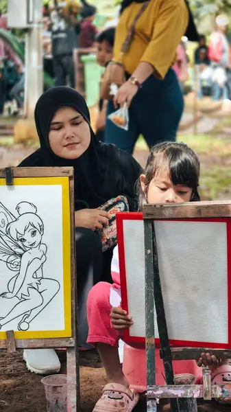 Bogor, Endonezya - 20 Nisan 2025: Müslüman bir anne yaratıcı bir açık hava sanat oturumu sırasında kızının resim yapmasını dikkatle izliyor