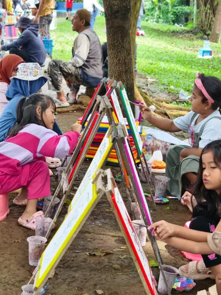 Bogor, Endonezya - 20 Nisan 2025: Çocuklar açık hava parkı ortamında renkli fırça ve sehpalarla yerde oturuyorlar. Eğitim, sanat içeriği ve ebeveynlik blogları için harika