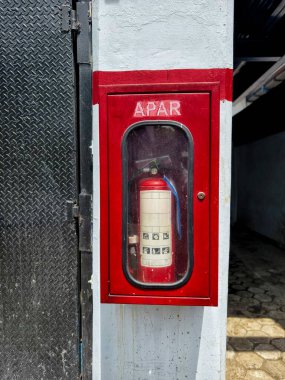 'APAR' etiketli koruyucu bir cam kutuya sarılı kırmızı bir yangın söndürücü yangın güvenliğine uyması için endüstriyel bir duvara monte edilmiş..