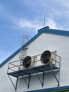 Dış duvarda havalandırma vantilatörleri ve güvenlik merdiveni bulunan sanayi binası, hava dolaşımı ve bakım girişi için gerekli.