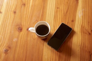 Ahşap bir masada dinlenmek, bir kahve fincanı ve akıllı telefon rahat, üretken bir atmosfer yaratır. Sabah rutinleri ya da evde ya da ofiste mola vermek için mükemmel bir ortam.