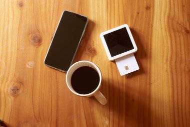 Temassız ödemeler, bir kredi kartı, akıllı telefon ve pos terminali kullanılarak, ahşap bir masada bir fincan kahvenin keyfini çıkarırken, kullanışlı ve modern bir ödeme deneyimi sunar.