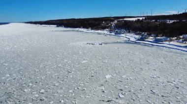 St. Lawrence nehrinin kıyılarında ve kıyılarında sıkıştırılan donuk yüzen buz şeridinin panoramik hava görüntüsü. Arka planda mavi bir gökyüzünün altında birkaç rüzgar türbini var..