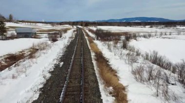 Ufukta bir dağ, kış mevsiminde körelmiş, karlı bir kırsal araziyi geçen bir tren yolu üzerinde düşük seviyeli uçuş..