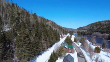 Kışın mavi bir gökyüzü altında iki dağ arasında akan somon nehri boyunca uzanan demiryolunu geçen karlı bir kırsal şeritteki eski evlerin panoramik manzarası. Routhierville, Quebec, Kanada, 2025.