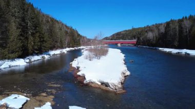 Tarihi Routhierville Kapalı Köprüsü 'ne bakan Matapedia Nehir yatağı üzerinde kışın karla kaplı bir dağ vadisinin zeminine karşı alçak uçuş. Quebec, Kanada, 2025.