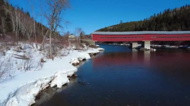 Matapdia Nehri üzerinde, tarihi Routhierville köprüsünün ahşap yapısının altından geçen bir uçuş, kışın arka planı olarak karla kaplı bir dağ vadisi. Quebec, Kanada, 2025.