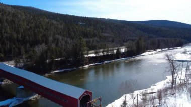 Karla kaplı bir dağ vadisinde nehri geçen eski kırmızı ahşap köprünün yukarıdan aşağı görüntüsü. Matapedia Nehri, Routhierville, Quebec, Kanada, 2025.