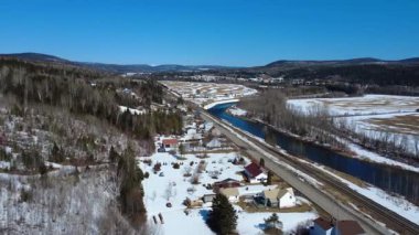 Dağlık bir vadide kıvrımlı bir nehrin yanında evleri, bir nehri ve bir demiryolu bulunan karla kaplı kırsal bir tarımın panoramik manzarası. Sainte-Florence, Quebec, Kanada, 2025.