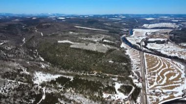 Karla kaplı bir dağ vadisinin panoramik yüksek irtifa manzarası bir nehir ve bulutsuz mavi gökyüzünün altında dolambaçlı bir yol ile kesişiyor. Matapedia Vadisi, Quebec, Kanada, 2025.