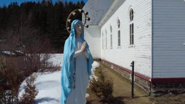 Kutsanmış Bakire Meryem 'in heykellerinden bir anıt. Elleri birbirine kenetlenmiş, bir tespih ve üç melek bir arada dua ediyor. Bir kilise ve yan camları yanında. Sainte-Florence, Quebec, Kanada, 2025.