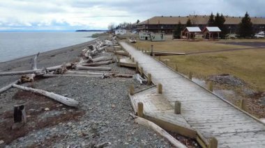 Tahta tahtalar ve küçük kazıklardan oluşan bir tahta kaldırım, baharın solmuş renklerinde bir halk parkının yakınındaki kayalar ve sürüklenen dallarla dolu bir sahil boyunca uzanır. Maria, Quebec, Kanada, 2025.