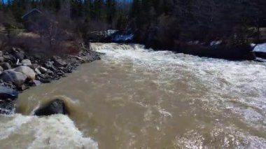 Kahverengi bir nehri kapsayan bir köprünün altından geçen bir dronun videosu kulübenin yakınındaki bir şelaleye doğru yükseliyor ve mavi gökyüzü altındaki bir rüzgar çiftliğini ortaya çıkarıyor, Quebec, Kanada, 2025.
