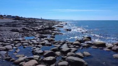 St. Lawrence Nehri kıyı şeridinde uçarken, dalgalar ve kayalar arasında güneşin yansımalarının parıldadığı yerde, mavi bir okyanus ve ufukta uzak bir kilise kulesi ile. Baie-des-Sables, Quebec 'te. Kanada, 2025.