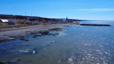 Eski kayalık rıhtımı, yüksek kilisesi ve mavi gökyüzünün altında bir rüzgar çiftliği olan büyüleyici küçük bir sahil köyünün kıyı şeridi manzarası. Baie-des-Sables, Quebec, Kanada, 2025.