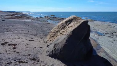 Dengesiz bir kaya parçası, deniz seviyesinin düşük olduğu görülen tortulu kayaların önündeki kumlu bir sahilde uzanır. Mavi okyanus ufukta gözüktüğü kadar uzanır. Baie-des-Sables, Quebec, Kanada, 2025.